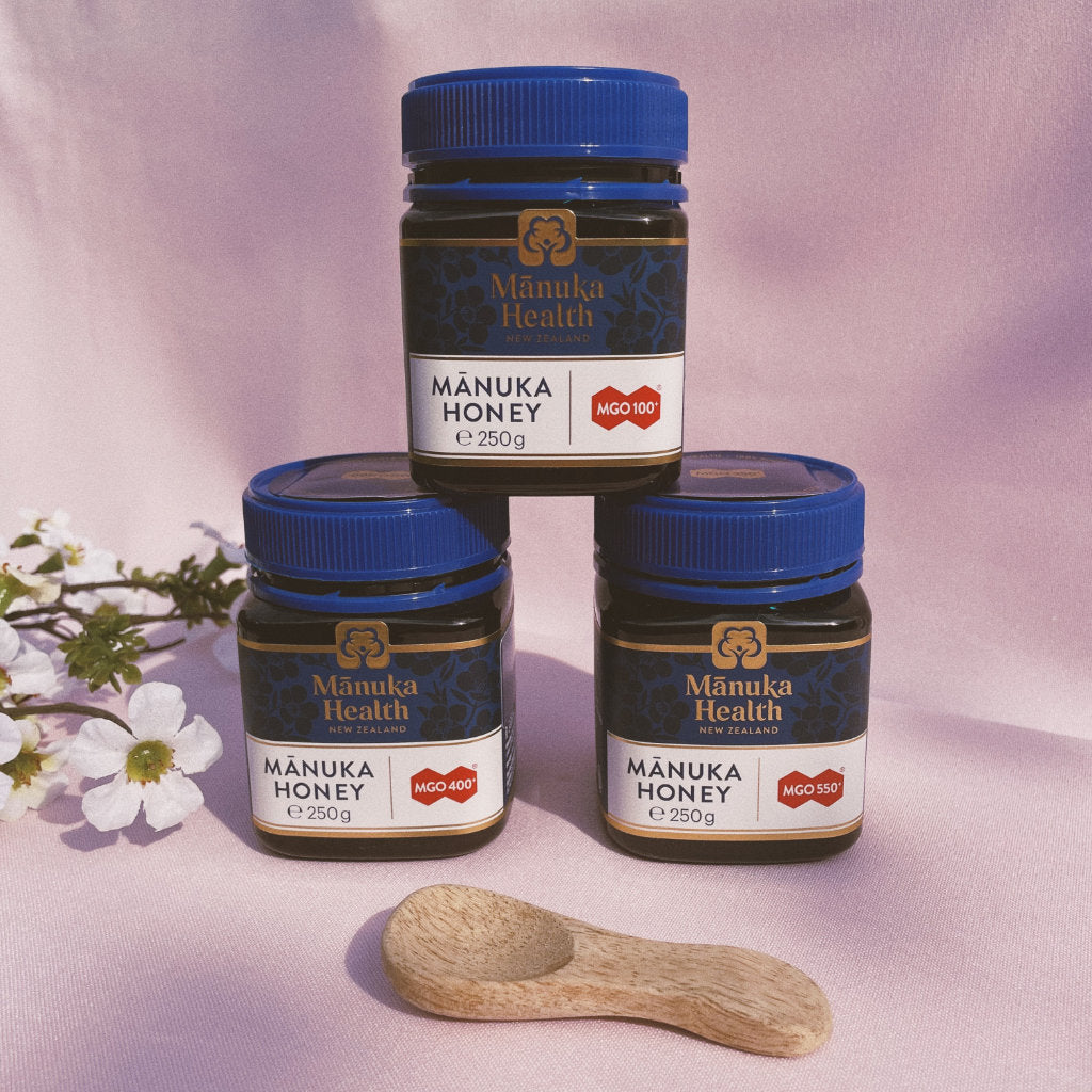 3 jars of Manuka honey by Manuka Health, on a pink background with a wooden spoon and a Manuka flower