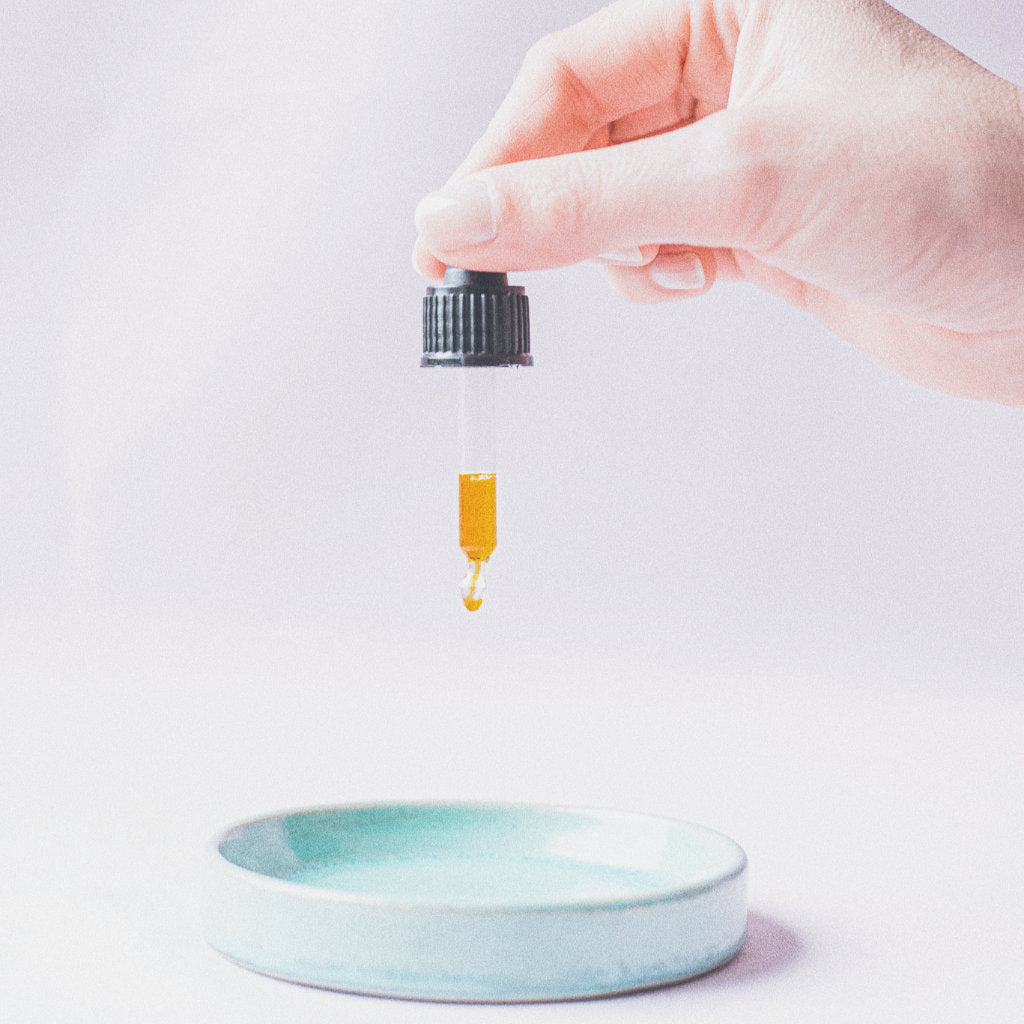 Hand holding a pipette with the Akasha blends Facial oil with golden colour above a turquoise plate